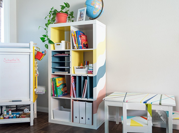 White IKEA cubby shelving with yellow and blue inserts, books, paper trays and school supplies beside a children's activity table